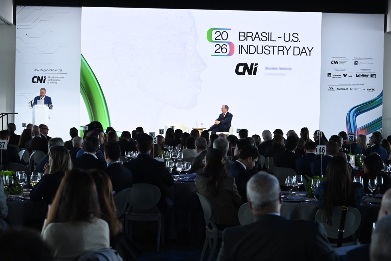 Auditório com mesas, público e grande tela com a marca Brasil‑U.S. Industry Day ao fundo.