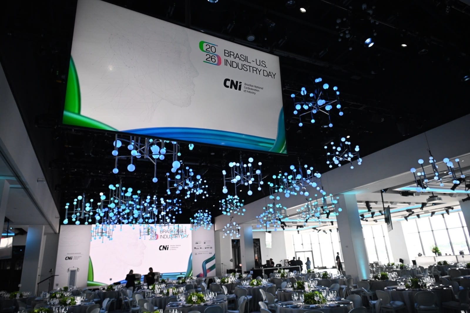 Salão de eventos com mesas postas, teto com elementos luminosos e tela grande ao fundo.