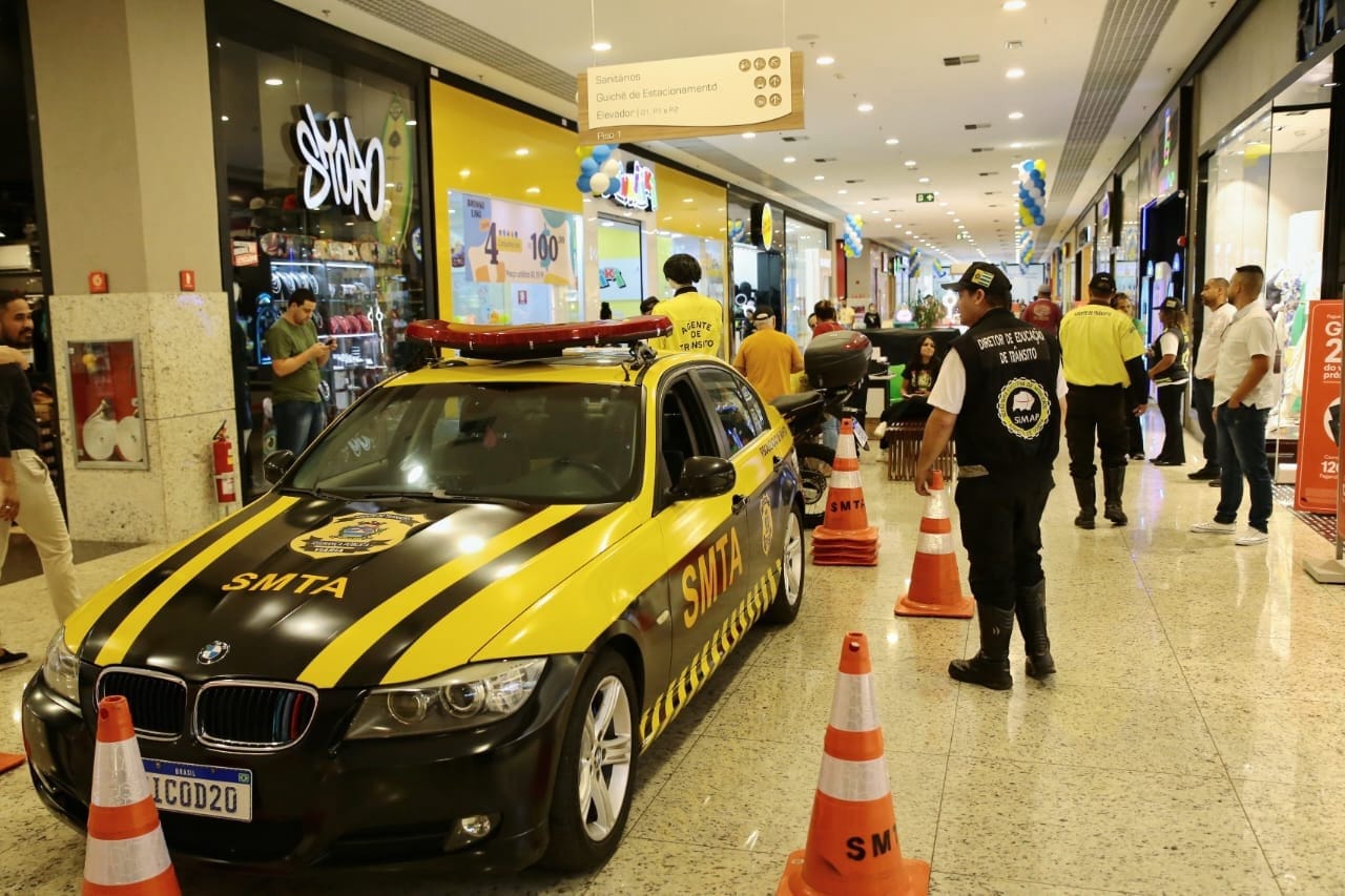 Viatura e motocicleta da SMTA posicionadas no corredor do shopping com cones; agentes em aproximação ao equipamento exposto