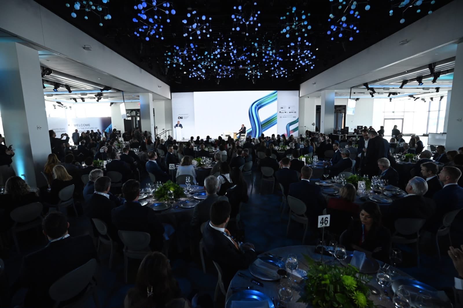Salão amplo com mesas redondas e convidados voltados para o palco principal.