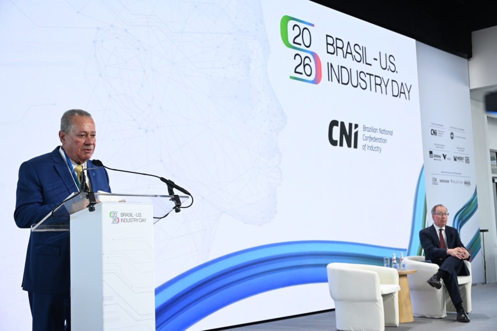 Cena lateral do palco com palestrante no púlpito e cadeira vazia ao lado ocupada por moderador.