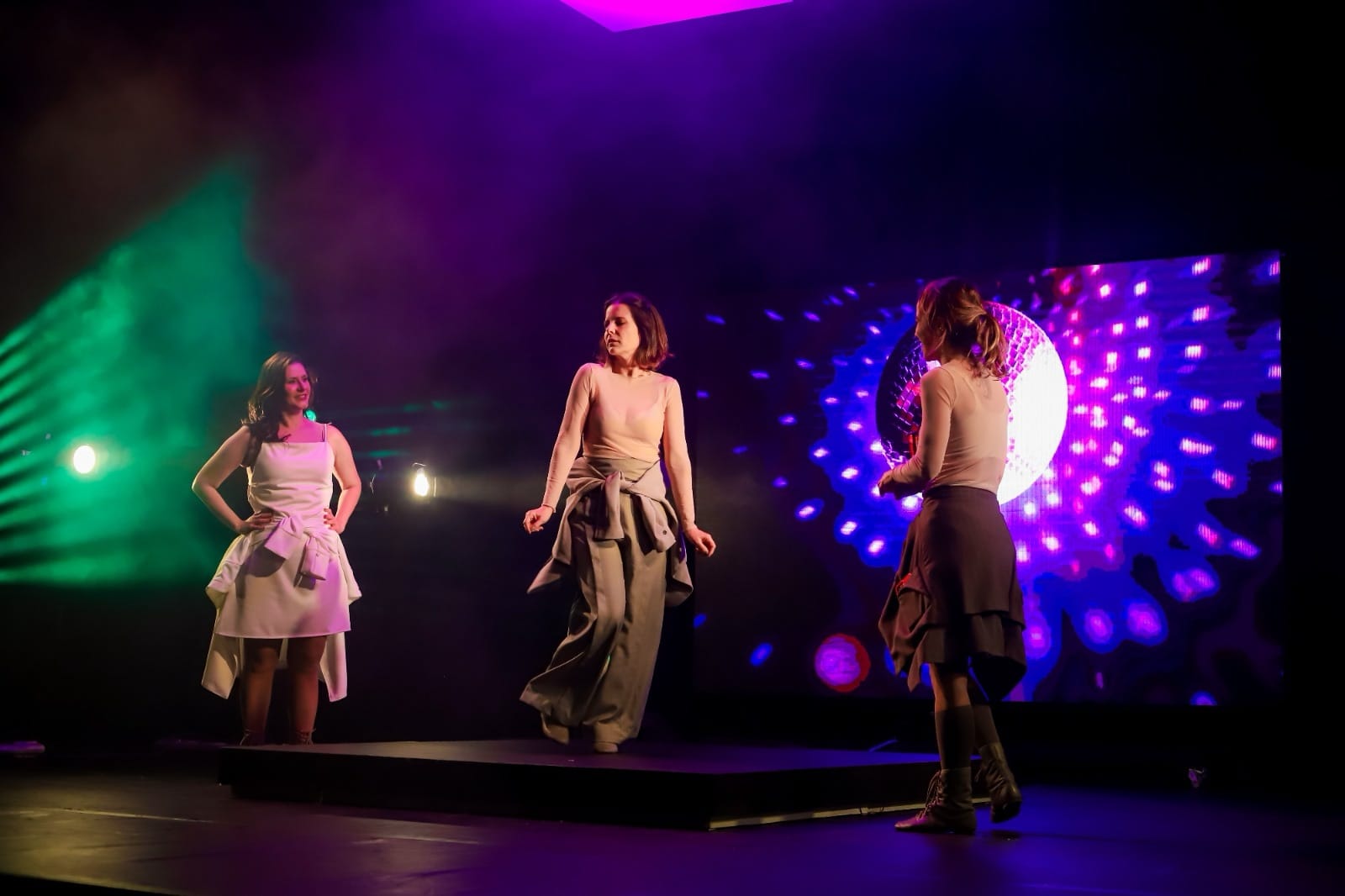 Cena do palco com três atrizes sob iluminação roxa e projeções ao fundo