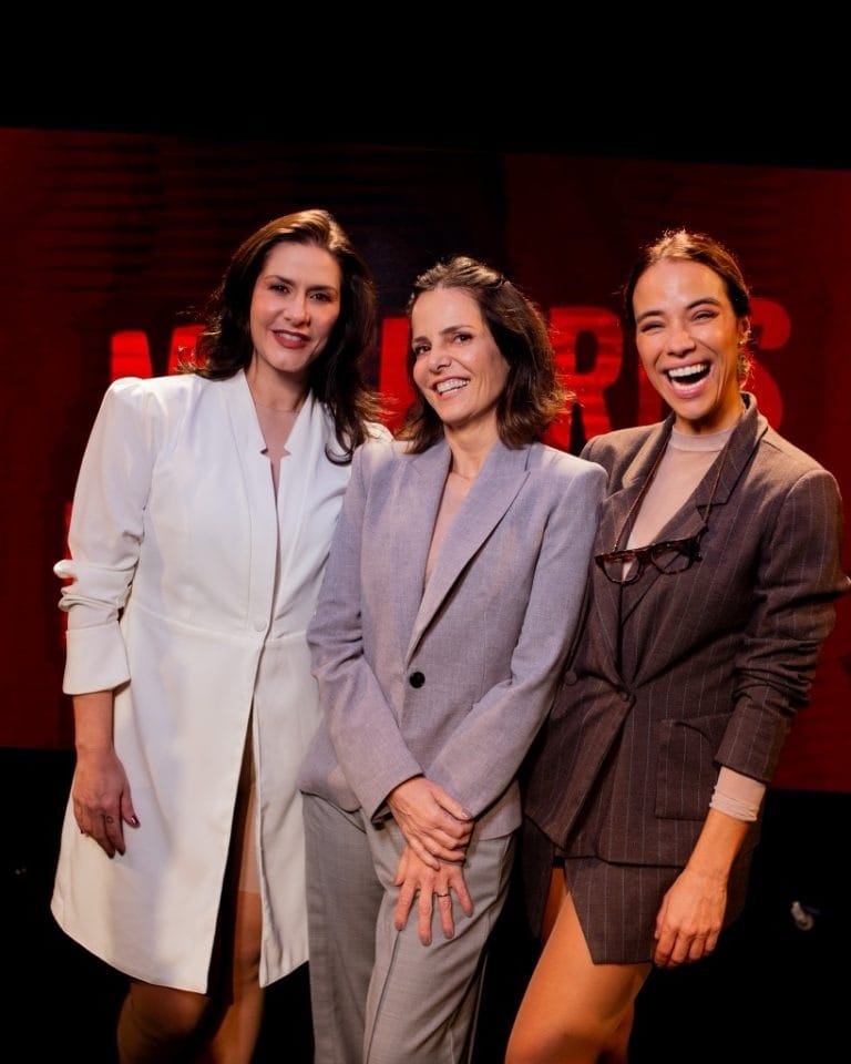 Três atrizes posando juntas nos bastidores, sorrindo
