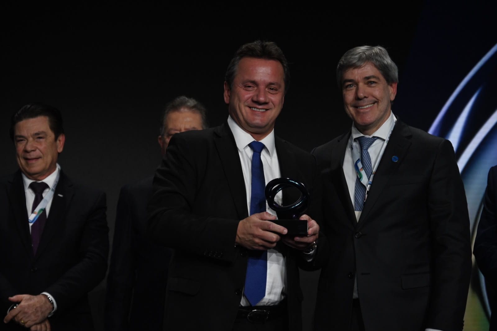 Dois executivos em traje formal seguram um troféu no palco; outros participantes ao fundo.
