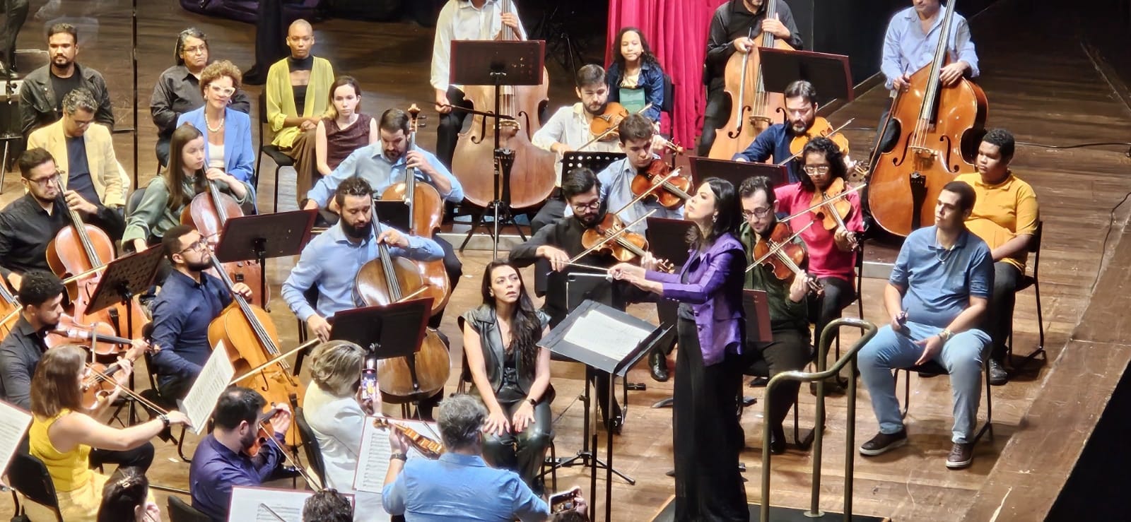 Regente em destaque conduzindo a orquestra, com músicos concentrados nas partituras