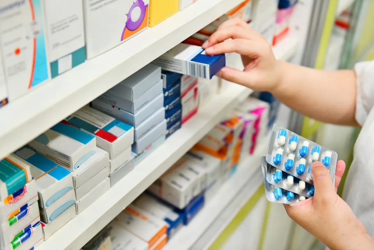 Prateleira de farmácia com caixas de medicamentos e mãos segurando cartelas de comprimidos.