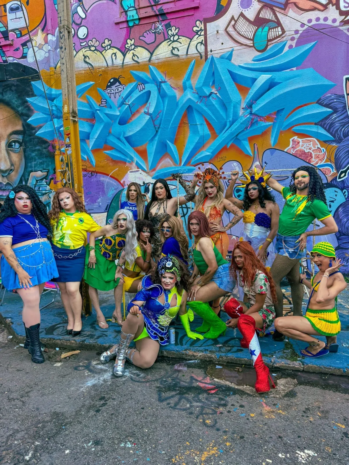 Grupo do Coletivo Drags Goiás em trajes com referências à seleção brasileira, posando em frente a um painel de grafite colorido