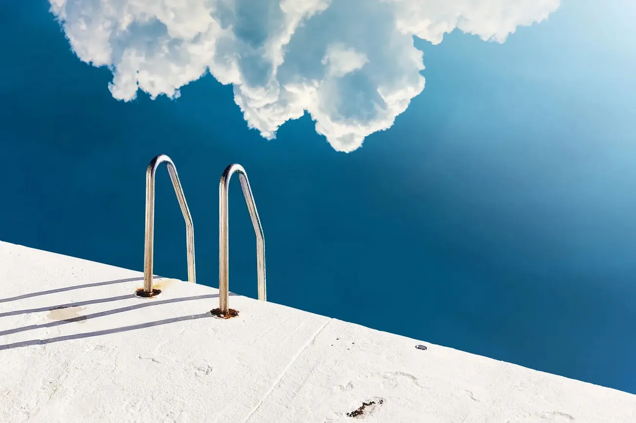 Borda de piscina branca com degraus metálicos e reflexo de nuvens sobre água azul intensa
