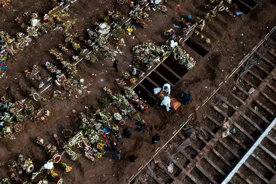 Vista aérea de sepulturas abertas e pessoas participando de enterro no cemitério, com flores e coroas ao redor das covas