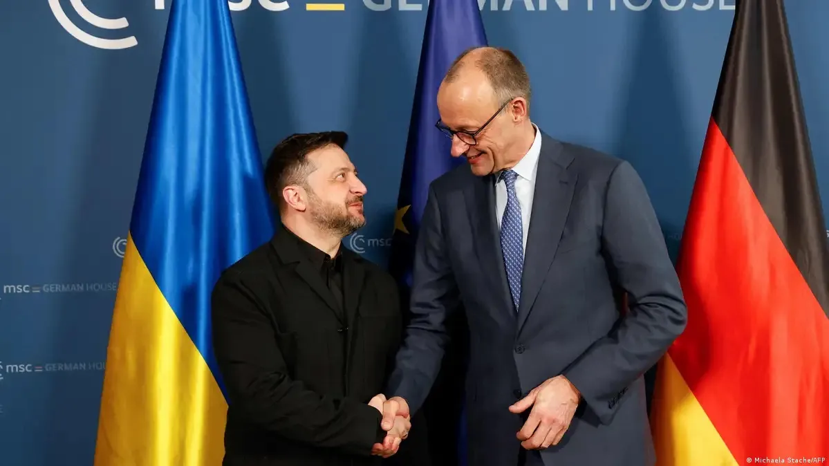 Volodimir Zelenski e o chanceler alemão Friedrich Merz apertando as mãos, com bandeiras da Ucrânia, União Europeia e Alemanha ao fundo