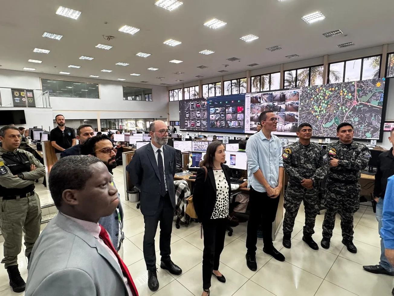 Comitiva e agentes em sala de comando de complexo prisional com painéis de monitoramento