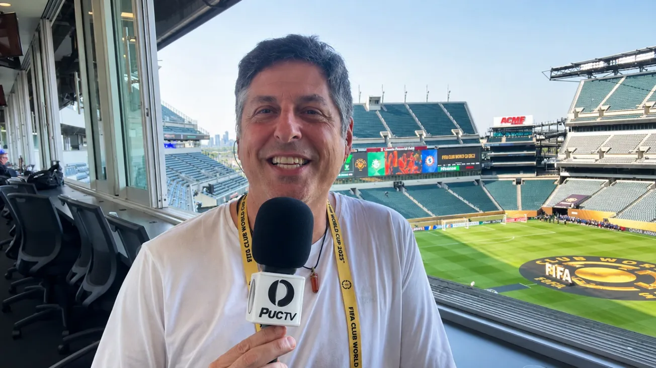 Leonardo Baran, correspondente da PUC TV, com microfone da emissora em cabine de imprensa de estádio