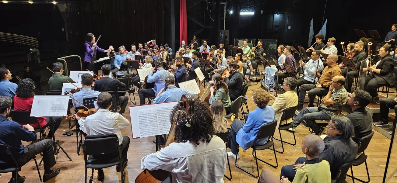 Orquestra filarmônica em palco com maestrina regendo, músicos de cordas e sopros em apresentação