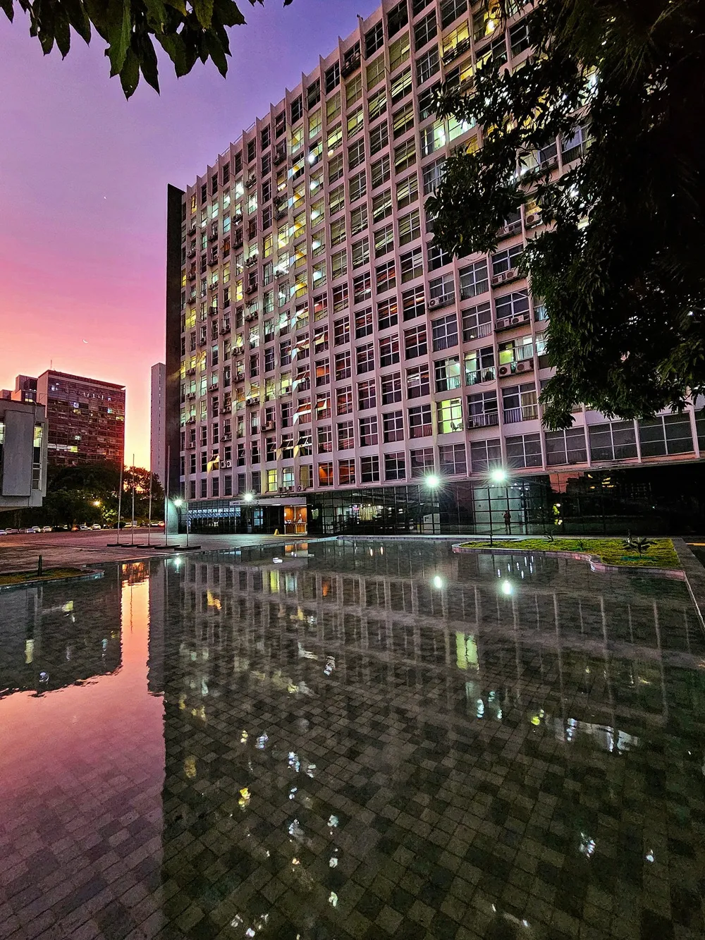 Fachada de edifício alto refletida em espelho d'água ao entardecer, com céu em tons rosados