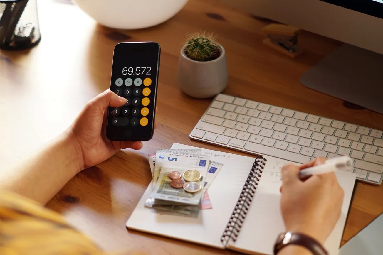 Pessoa calculando despesas com celular e caderno; sala de atendimento do NAF; mãos usando calculadora sobre documentos