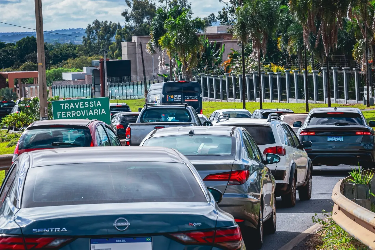 Fila de carros no trânsito com placa indicativa 'Travessia Ferroviária'; vista traseira de veículos em via urbana